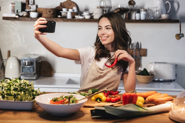 Hermosa joven vistiendo delantal cocinar ensalada saludable en la cocina de casa, tomando un selfie con teléfono móvil