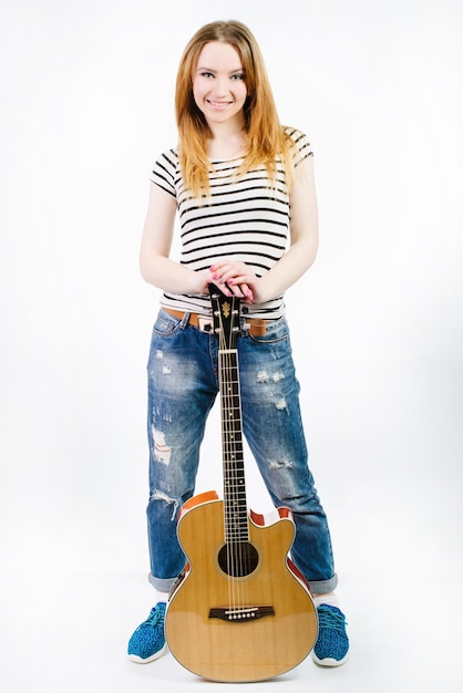 Hermosa joven vistiendo jeans y una camiseta a rayas con una guitarra acústica sobre un fondo blanco.