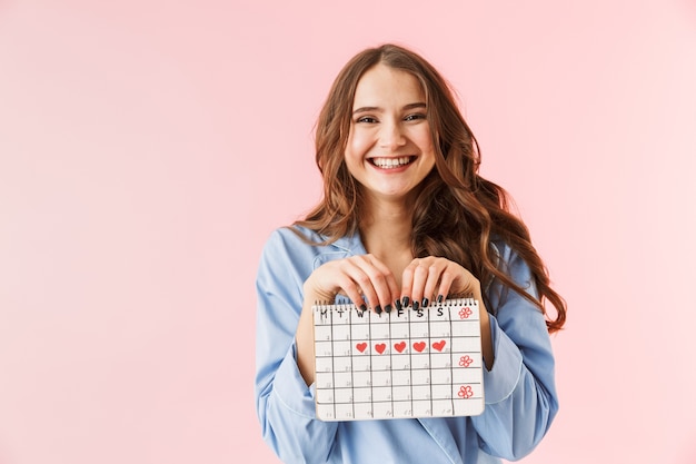 Hermosa joven vistiendo pijamas que se encuentran aisladas sobre fondo rosa, mostrando el calendario menstrual