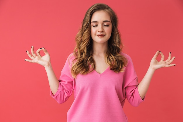 Hermosa joven vistiendo ropa casual que se encuentran aisladas sobre pared rosa, meditando