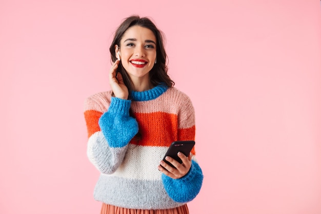 Hermosa joven vistiendo ropas coloridas que se encuentran aisladas sobre rosa, escuchando música con auriculares, sosteniendo el teléfono móvil