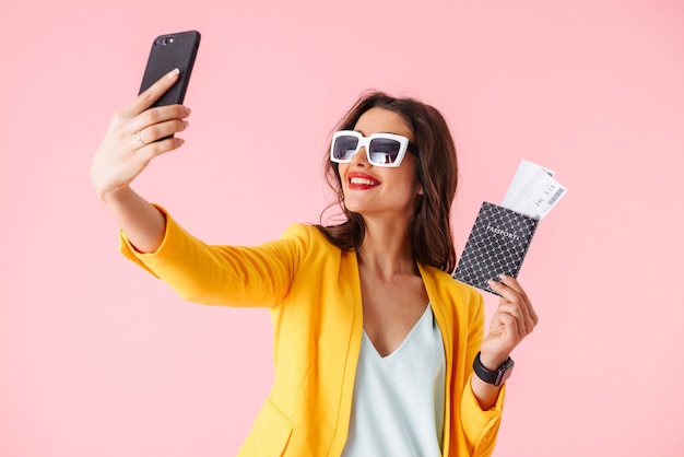 Hermosa joven vistiendo ropas coloridas que se encuentran aisladas sobre rosa, mostrando el pasaporte con boletos de avión, tomando un selfie