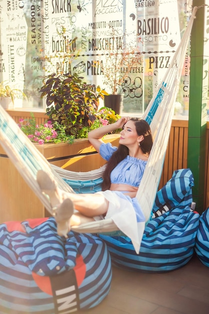 Una hermosa joven yace en una hamaca en la terraza de verano de un café de la ciudad y disfruta del verano