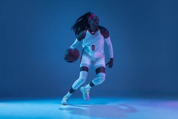 Hermosa jugadora de baloncesto afroamericana en movimiento y acción en luz de neón en la pared azul concepto de afición de deporte profesional de estilo de vida saludable