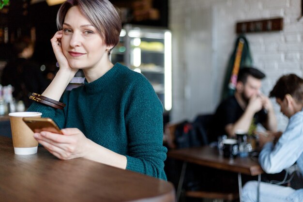 Hermosa linda chica en el café mirando a la cámara con café sonriendo. Jersey de punto verde. chica vestida a la moda. chica mira el teléfono. dama de negocios