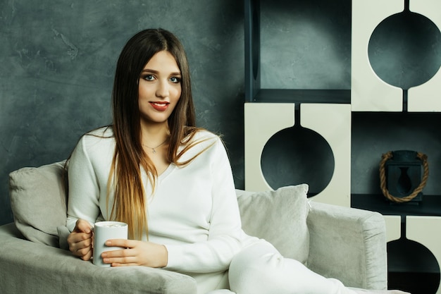 Hermosa y linda Hermosa joven sosteniendo una taza de café y sentada en una silla en casa Concepto de estilo de vida