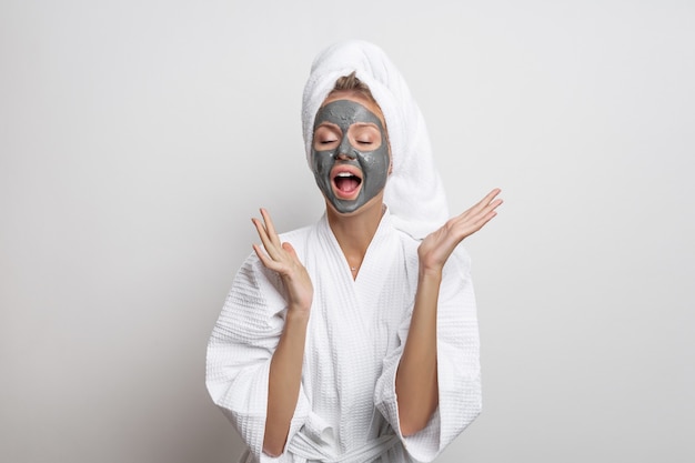 Hermosa linda modelo posando en una bata de baño de gofre blanco y una toalla en la cabeza posando con las manos separadas y la boca abierta con una máscara de arcilla en el rostro sobre un fondo blanco.