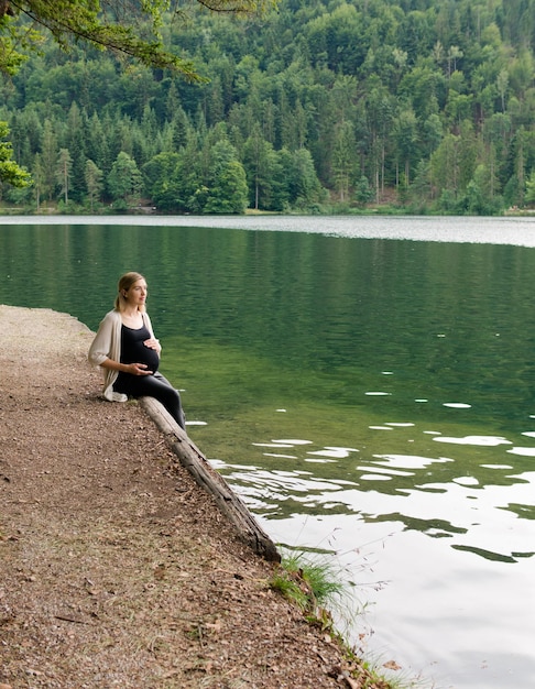 Hermosa y linda mujer embarazada se sienta cerca de un lago de montaña en un bosque en la orilla