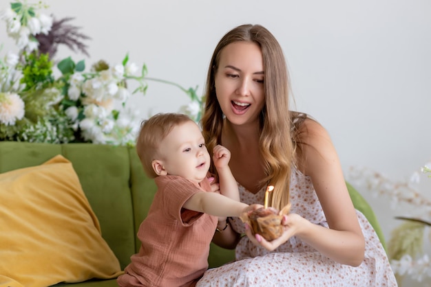 Hermosa madre amorosa con un vestido se sienta junto a su pequeño hijo en un sofá verde rodeado de hermosas flores Sostiene un pastelito con una vela en la mano y se regocija Postre festivo