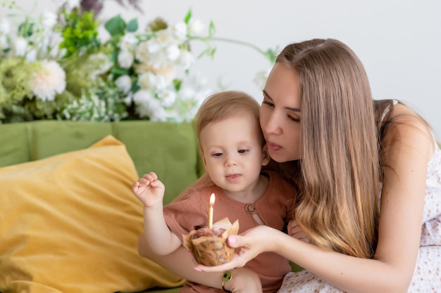 Hermosa madre amorosa con un vestido se sienta junto con su pequeño hijo en un sofá verde rodeado de hermosas flores Sostiene un pastelito con una vela en la mano Sopla la vela Postre festivo