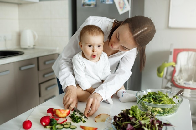 Hermosa madre de cabello oscuro cocinando la cena frente a su adorable hijo. Niño mirando a un lado con expresión desconcertada.