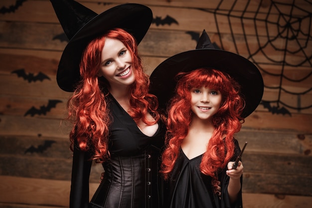 Foto hermosa madre caucásica y su hija con pelo largo rojo en trajes de bruja y varita mágica celebrando la presentación de halloween