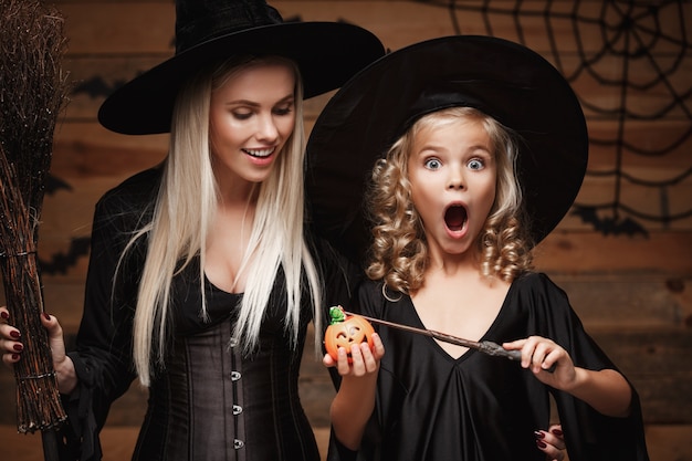 hermosa madre caucásica y su hija en trajes de bruja disfrutan el uso de magia con varita mágica para el tarro de calabaza de halloween