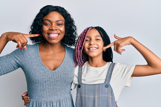 Hermosa madre e hija afroamericanas vestidas con ropa informal y abrazándose sonriendo alegremente mostrando y señalando con los dedos los dientes y la boca. concepto de salud dental.