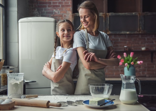 Foto hermosa madre e hija en delantales se miran y sonríen mientras están de espaldas en la cocina en casa