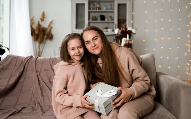 Hermosa madre e hija están sentadas en el sofá con un regalo y mirando a la cámara