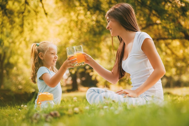 Hermosa madre e hija en el parque haciendo un pequeño picnic. Están disfrutando y bebiendo jugo de naranja.