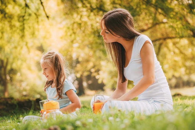 Hermosa madre e hija en el parque haciendo un pequeño picnic. Están disfrutando y bebiendo jugo de naranja.