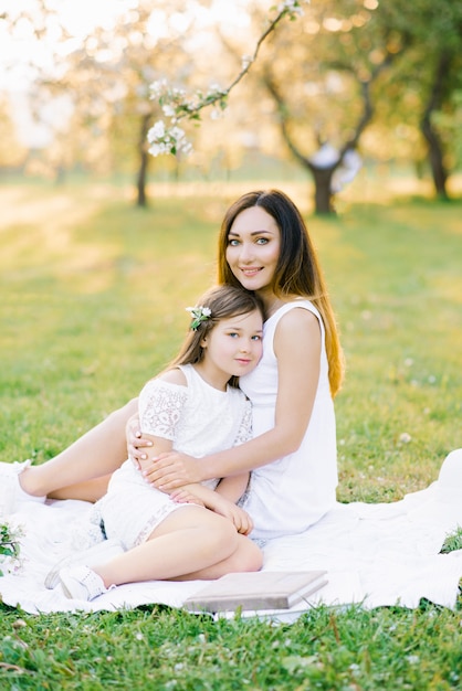 Hermosa madre de familia y su hija en vestidos blancos sentados en una tela escocesa blanca en los jardines de flores en la primavera