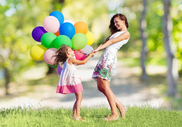 Hermosa madre feliz con hija divirtiéndose en campo verde sosteniendo globos de colores