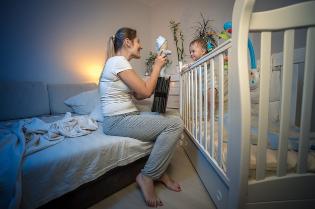 Hermosa madre feliz jugando con su bebé en la cuna por la noche