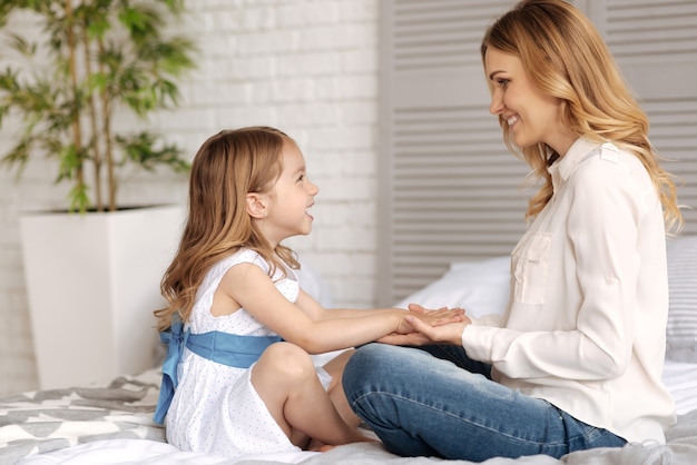 Hermosa madre feliz sosteniendo las manos de su pequeña hija, que está vuelta hacia abajo en su palma, mientras ambos se sonríen el uno al otro