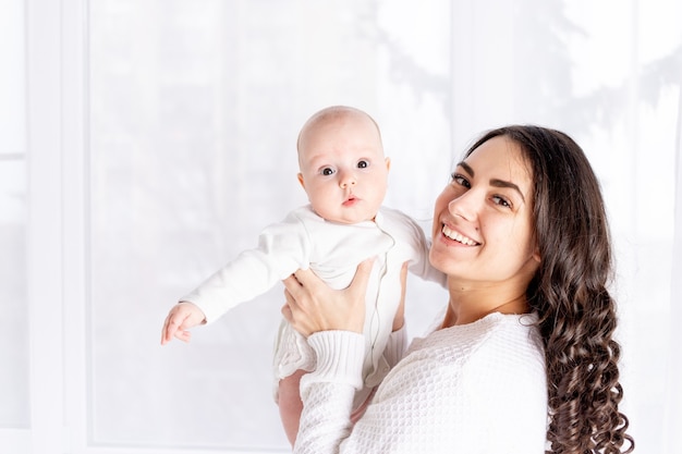 Hermosa madre sosteniendo a un bebé en sus brazos en la ventana de la casa, el concepto de una familia amorosa feliz, estilo de vida