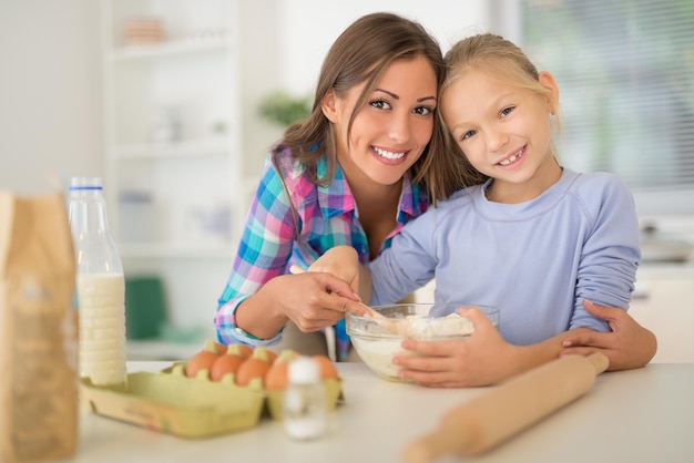 Hermosa madre y su hija haciendo masa juntos en la cocina doméstica. Mirando a la cámara.