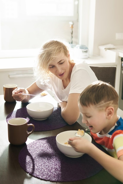 Hermosa madre y su lindo hijo comiendo copos de maíz saludables para el desayuno en la mañana soleada