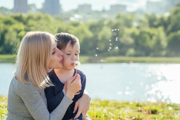 Hermosa madre y su lindo hijo soplando un globo de diente de león en el parque sobre un fondo de hierba verde, árboles y lago. el concepto de vacaciones familiares en la naturaleza.