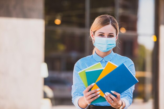 La hermosa maestra con una máscara médica muestra libros coloridos al aire libre