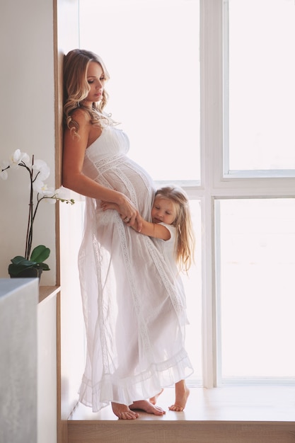 Hermosa mamá embarazada con niño posando junto a la ventana en casa