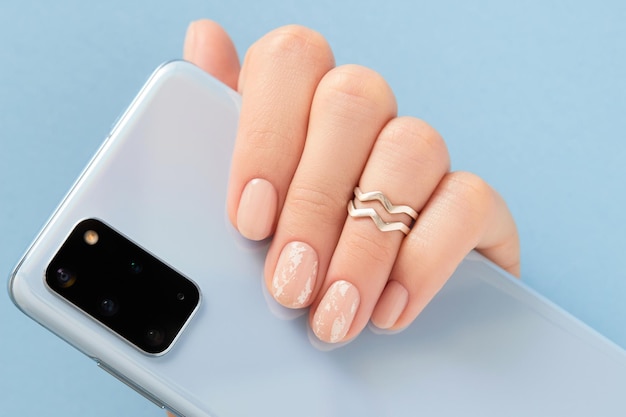 Hermosa mano de mujer con diseño de uñas desnuda sosteniendo el teléfono inteligente sobre fondo azul manicura pedicura