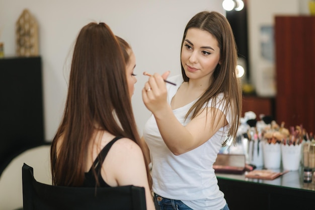 Hermosa maquilladora maquillando a una joven pelirroja en un salón de belleza sentada frente a un gran espejo Concepto de preparación para las vacaciones y la reunión