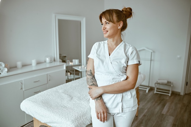 Hermosa masajista mujer caucásica feliz está posando en el salón de tratamiento moderno minimalista blanco