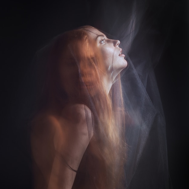 Foto hermosa modelo con cabello rojo posando detrás de una tela negra y un velo en un estudio.