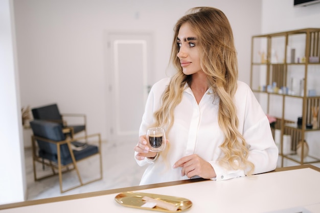 Hermosa modelo esperando el procedimiento de cosmetología y bebiendo café en el vestíbulo interior blanco de belleza