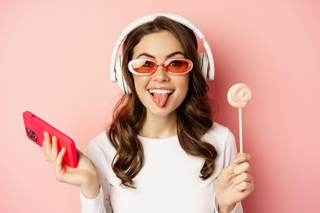 Hermosa modelo femenina escuchando música en auriculares, sosteniendo lolipop y teléfono móvil, posando con gafas de sol, de pie sobre fondo rosa.