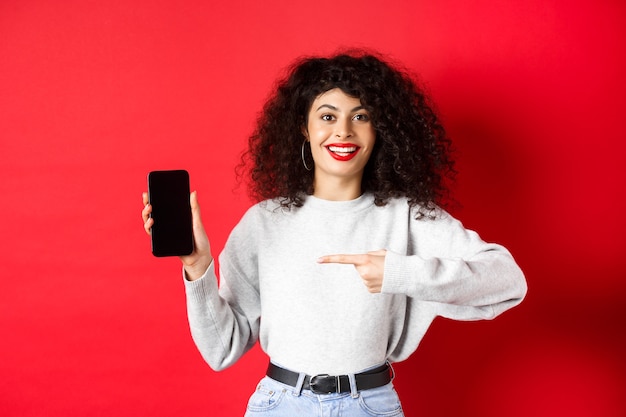 Hermosa modelo femenina que muestra la pantalla del teléfono inteligente vacía, apuntando a la pantalla del teléfono y sonriendo, de pie contra el fondo rojo