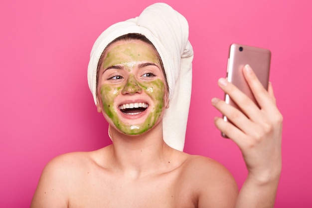 Hermosa modelo femenina tiene una máscara de pepino en la cara, envuelta en una toalla blanca, posa semidesnuda, toma selfie en el teléfono inteligente, posa con una sonrisa con dientes, aislado en rosa. Concepto de cosmetología.