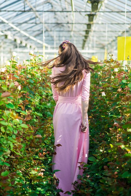 Hermosa modelo femenina vestida con un largo vestido rosa posa entre las muchas flores en el invernadero