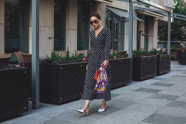 Hermosa modelo mira mujer morena con un vestido blanco con lunares negros es mientras se despierta en un fondo de calle de la ciudad con elegantes bolsos en la mano.