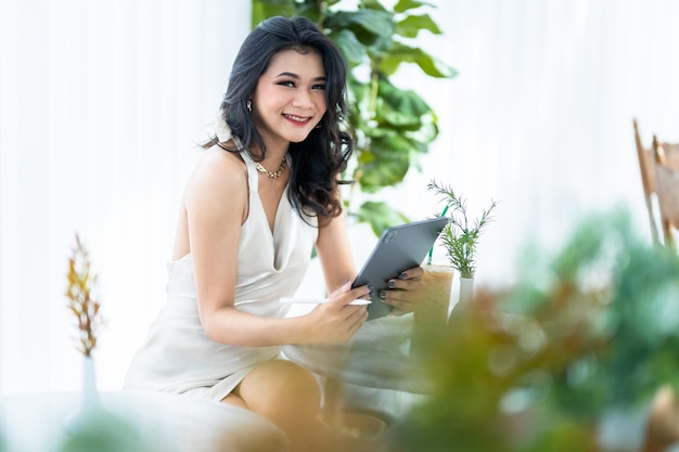 Hermosa modelo de moda de mujer de negocios de éxito independiente asiática en vestido blanco sin mangas de verano usando un trabajo informal con tableta para navegar por Internet, chatear y bloguear en una cafetería