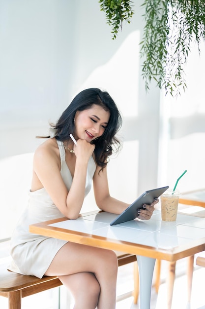 Hermosa modelo de moda de mujer de negocios de éxito independiente asiática en vestido blanco sin mangas de verano usando un trabajo informal con tableta para navegar por Internet, chatear y bloguear en una cafetería