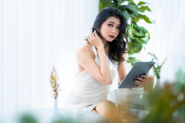 Hermosa modelo de moda de mujer de negocios de éxito independiente asiática en vestido blanco sin mangas de verano usando un trabajo informal con tableta para navegar por Internet, chatear y bloguear en una cafetería