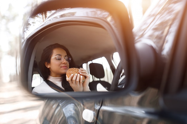 Foto hermosa modelo morena almuerza en un auto. mujer come una hamburguesa