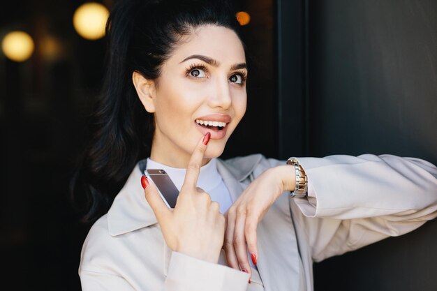 Hermosa modelo morena de ensueño con pestañas largas y rizadas, ojos oscuros y brillantes y manicura roja vestida con una chaqueta blanca sosteniendo su dedo en los labios mirando hacia arriba mientras sueña con algo agradable