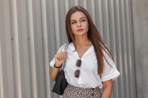 Hermosa modelo de mujer de negocios de moda con ropa informal de verano con una elegante camisa blanca y falda con gafas de sol antiguas y un bolso de mano camina por la calle