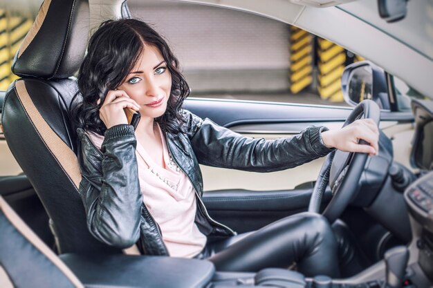 Hermosa modelo de mujer sexy en ropa de cuero con un coche blanco y un teléfono inteligente en el estacionamiento