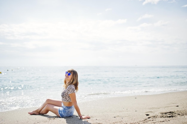 Hermosa modelo relajándose en una playa de mar usando jeans, camisa corta de leopardo y gafas de sol